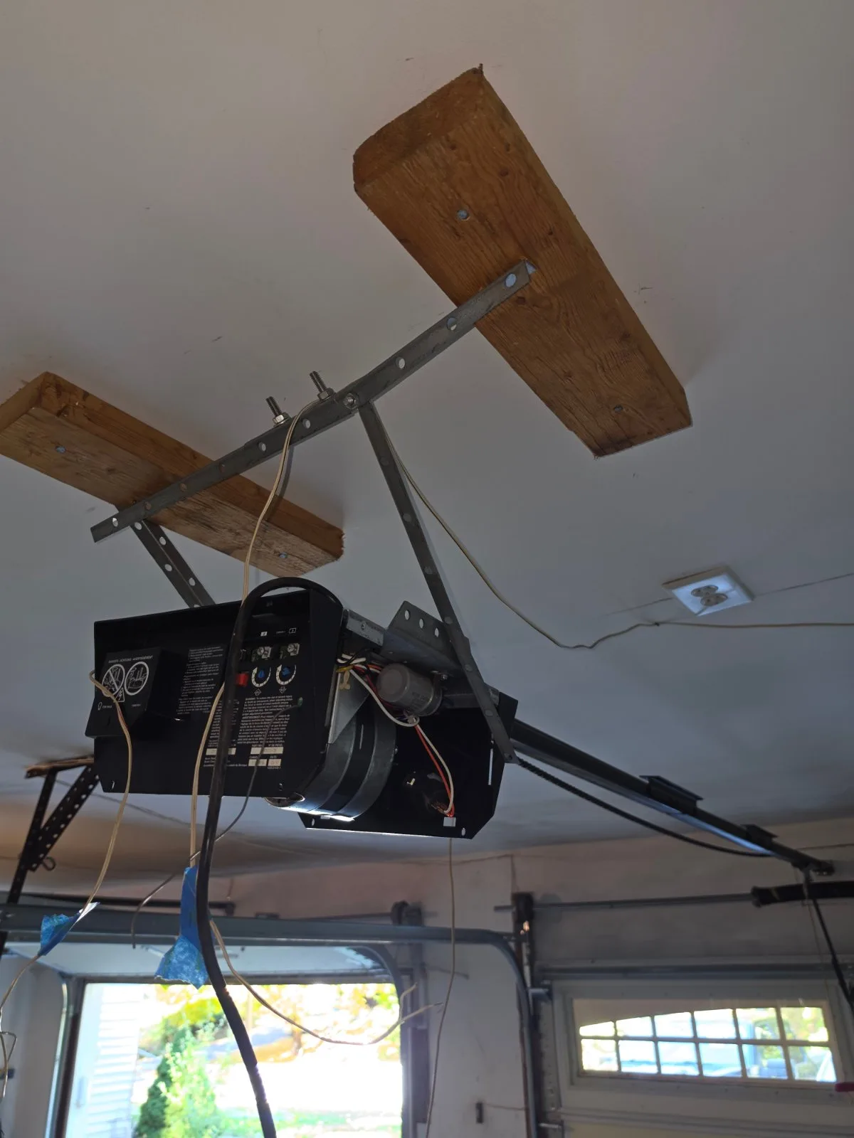 An older garage door opener motor with exposed wiring mounted to wooden blocks on the ceiling of a garage.